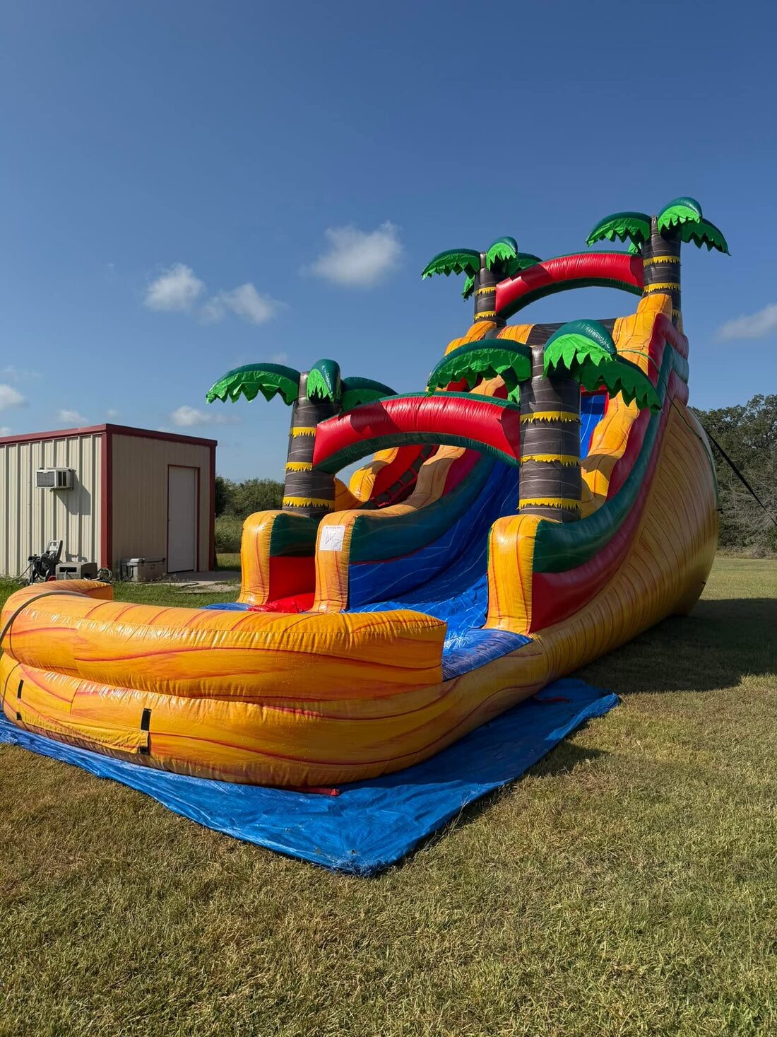 Tall orange and yellow tropical water slide with palm trees, set up on grass under a blue sky