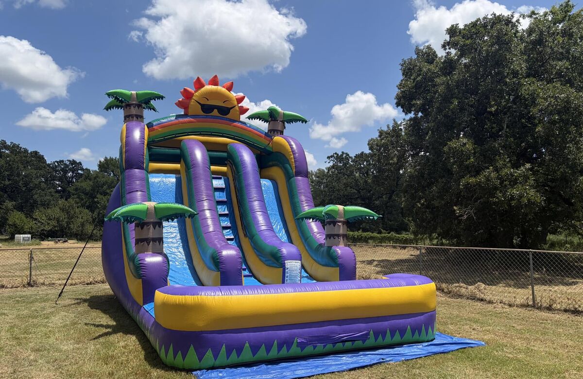 Purple and green water slide with palm trees and a sun on top, set up in a field